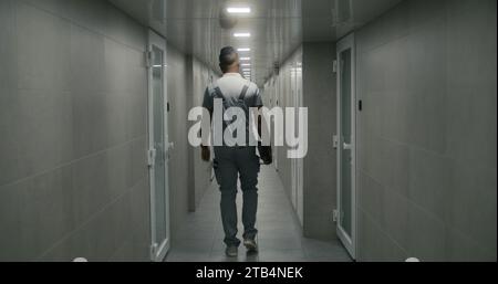 African American professional engineer in uniform walks in corridor of modern factory or plant holding tablet computer. Heavy industry worker in hallway of office building at industrial facility. Stock Photo