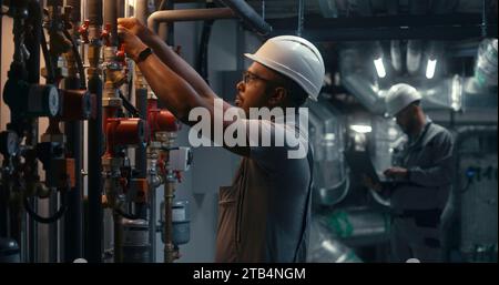 African American heavy industry worker wearing safety uniform and hard hat inspects piping system on modern plant or industrial energy facility. Professional engineer maintains manufacturing factory. Stock Photo