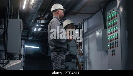 Professional Caucasian engineer in safety uniform, goggles and hard hat uses digital tablet computer, adjusts energy system on modern factory. Technician worker talks with African American colleague. Stock Photo