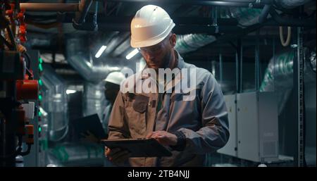 Diverse team of factory specialists work on modern plant using laptop and tablet computers. Industrial workers wearing safety uniform and hard hat inspect pipeline system. Heavy industry concept. Stock Photo