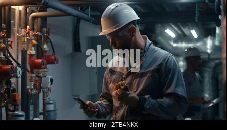 Professional engineer works at modern factory, eats burger and uses phone. In background African American colleague inspects pipe system using tablet computer. Industrial manufacturing plant concept. Stock Photo