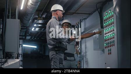 Professional Caucasian engineer in safety uniform, goggles and hard hat uses digital tablet computer, adjusts energy system on modern factory. Technician worker talks with African American colleague. Stock Photo