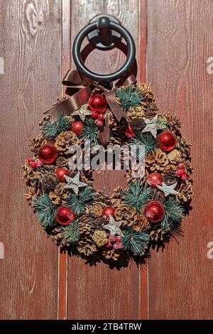 Rustic Christmas wreath hanging on wooden door. Wreath is decorated with natural pine cones, red baubles, and small green fir branches, and few silver Stock Photo