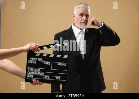 Senior actor performing role while second assistant camera holding clapperboard on beige background, selective focus. Film industry Stock Photo