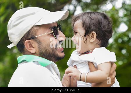 Happy Young indian father wearing hat and sunglasses lifting and playing with his daughter showing love and care in park or garden. Stock Photo
