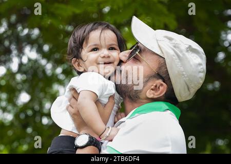 Happy Young indian father wearing hat and sunglasses lifting and kissing his daughter showing love and care in park or garden. Stock Photo
