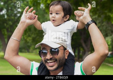 Happy Young indian father wearing hat and sunglasses holding hands and carry his daughter on back or shoulder having fun in park or garden. Stock Photo