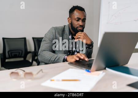 Serious black male programmer developer coding software on laptop computer typing on keyboard sitting at table in modern office room. Stock Photo