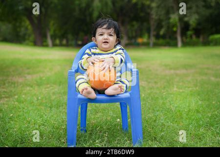 Cute baby boy sitting on blue chair in a garden playing with ball Stock Photo