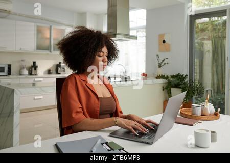Serious woman working on laptop at kitchen table at home Stock Photo