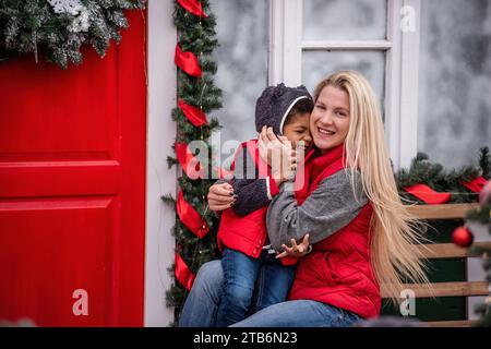 Caucasian mother with African American son having fun in backyard, on bench near red door with wreath. Young blonde woman kisses little boy, tickles h Stock Photo