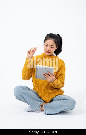 Beautiful and thoughtful Asian woman in casual clothes sits on an isolated white background and uses her digital tablet. People and technology concept Stock Photo