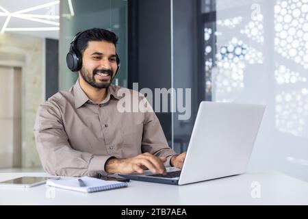 Young successful Indian programmer at workplace inside office, man with headphones smiling satisfied, coding new software using laptop at table, businessman in business shirt successful, satisfied Stock Photo