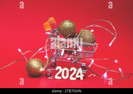 Christmas decoration tree toy balls in a small shopping trolley cart with garland and wooden numbers 2024 year on a red festive background. New Year, Stock Photo