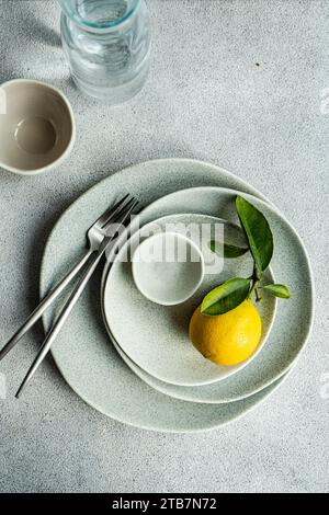 A vibrant lemon with leaves placed on a set of speckled ceramic plates, accompanied by sleek cutlery and glassware on a textured surface. Stock Photo