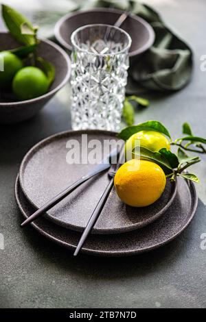 A sophisticated table setting featuring black cutlery on a dark plate with a bright yellow lemon, alongside a textured glass and fresh limes. Stock Photo