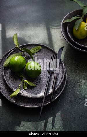 A sophisticated table setting featuring dark plates, cutlery, and fresh green limes on a moody, shadowed surface. Stock Photo