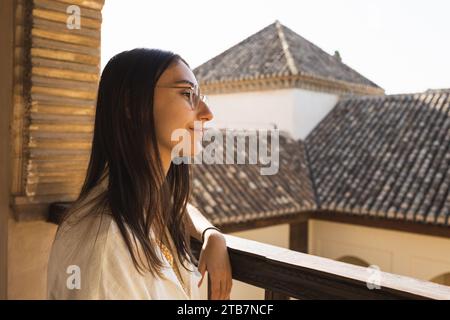 Side view of thoughtful smiling female tourist wearing eyeglasses and looking away while standing by wooden railing in historic place against house in Stock Photo