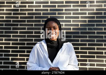 Portrait of confident smiling African American Muslim female entrepreneur in black hijab and white formal standing against brick wall in city Stock Photo