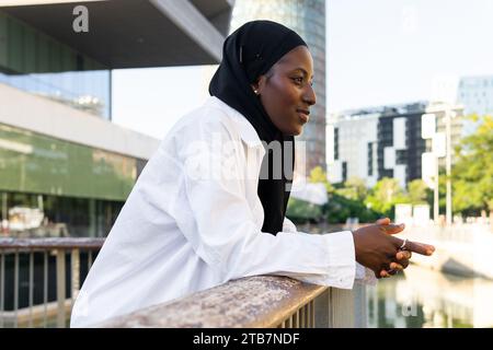 Side view of thoughtful African American Muslim businesswoman in black hijab looking away while leaning on railing in city Stock Photo