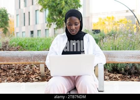 Serious concentrated Muslim female entrepreneur in traditional headscarf sitting on bench and using laptop while working on new business project Stock Photo