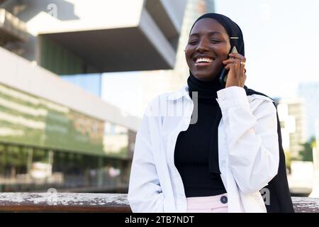 Cheerful African American young businesswoman in hijab with eyes closed talking over smart phone while standing against modern building Stock Photo