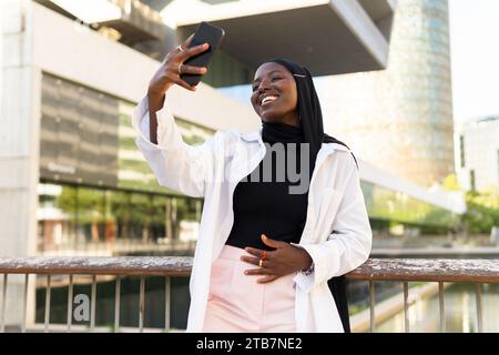 Cheerful Muslim female entrepreneur dressed in formals and black hijab taking selfie with mobile phone while standing by railing against modern buildi Stock Photo
