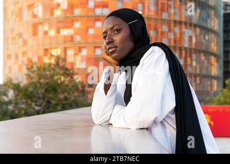 Side view of thoughtful African American Muslim businesswoman in black hijab touching chin and looking away while leaning on railing in city Stock Photo