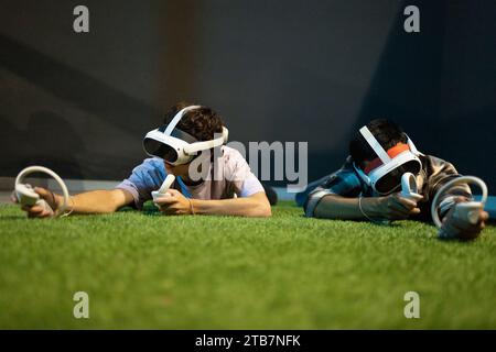 Two young individuals are immersed in a virtual reality experience, lying on their stomachs with VR headsets and controllers Stock Photo