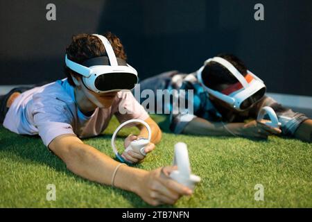 A young man lies prone on artificial grass, engaged in virtual reality gaming with a VR headset and handheld controllers Stock Photo