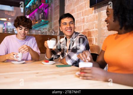 Cheerful multiethnic group of young adults engaged in conversation at a wooden table in a stylish cafe setting with a cup of coffee. Stock Photo