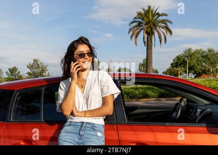 Happy attractive Caucasian young woman leaning on orange car and talking through on smartphone against sky on sunny day Stock Photo