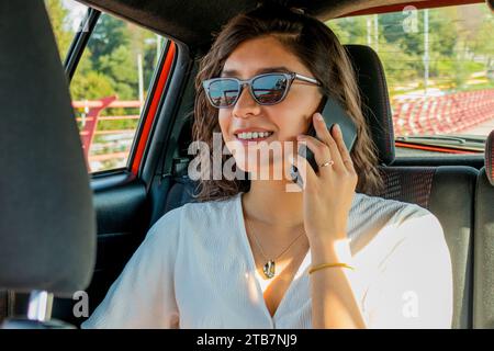 Smiling beautiful Caucasian young woman wearing sunglasses and casuals listening to smart phone during call while sitting in car backseat Stock Photo