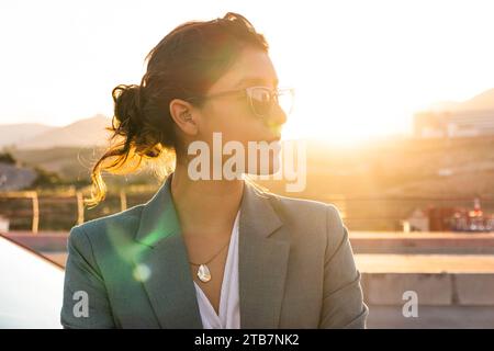 Serious Caucasian businesswoman wearing sunglasses and blazer looking away confidently while standing against sky with back lit in city during sunset Stock Photo