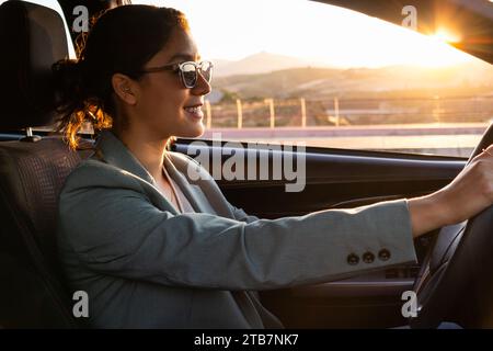 Portrait of cheerful young beautiful Caucasian executive wearing sunglasses and blazer driving a modern car at sunset in city Stock Photo