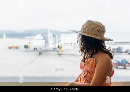 Side view of faceless girl traveler wearing straw hat and casuals looking at passenger airplane on taxiway through window at airport Stock Photo