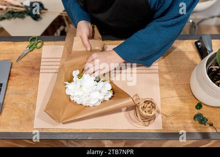 Top view of hands of unrecognizable person crafting a bouquet, wrapping white flowers in brown paper on a wooden table, with twine and scissors nearby Stock Photo