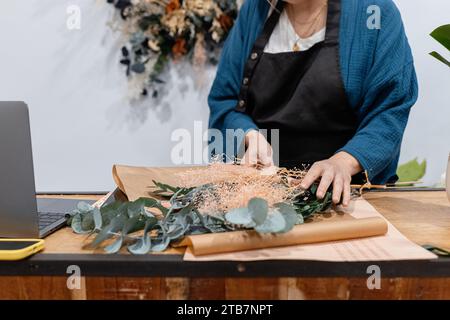 Anonymous florist meticulously arranges greenery for a bouquet on a table, with tools and laptop nearby. Stock Photo
