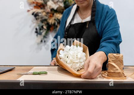 Unrecognizable person crafting a bouquet, wrapping white flowers in brown paper on a wooden table, with twine and scissors nearby Stock Photo