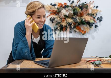 Smiling businesswoman florist on a phone call while working on her laptop at a wooden counter looking at screen, with flowers in the background Stock Photo