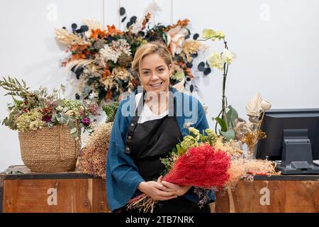 Smiling female florist standing looking at camera in flower shop, holding a bright, red bouquet, surrounded by a diversity of colorful floral arrangem Stock Photo