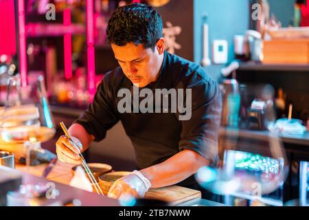 Focused male cook in black uniform and gloves with chopsticks and ingredients making fresh sushi in kitchen while working in illuminated nightclub Stock Photo