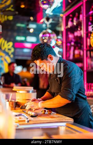 Side view of Confident young male chef with in black uniform and gloves standing while placing freshly cooked sushi on tray at kitchen at restaurant Stock Photo
