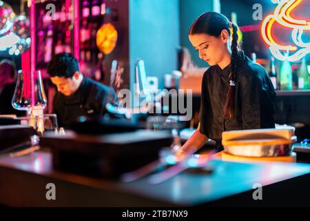 Beautiful focused young chef with braided hair cooking with male coworker in modern kitchen at illuminated sushi bar Stock Photo