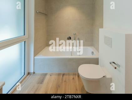 A contemporary bathroom featuring a tub, toilet and minimalist aesthetics, with ample natural light. Stock Photo