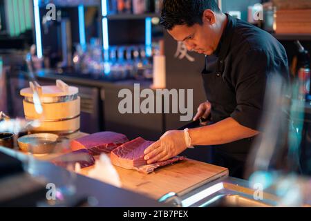 A sushi chef attentively slices fresh fish with a sharp knife at a wooden counter in the ambient lighting of a modern sushi restaurant. Stock Photo