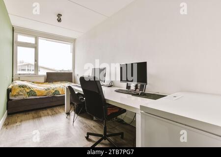 Computers on desk with empty chairs and small bed by bright window in white bedroom at modern apartment Stock Photo