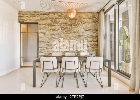 Stylish light hanging over dining table and chairs in dining room with brick wall and large window at modern home Stock Photo
