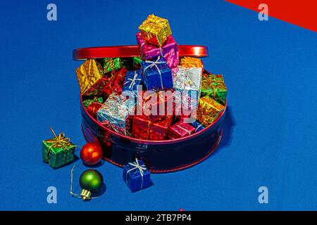 Top view red container brimming with brightly wrapped Christmas gifts on a blue surface with a red and blue backdrop Stock Photo