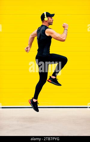 Side view full body of energetic male sportsman wearing headphones and black activewear jumping in air over street against yellow wall Stock Photo